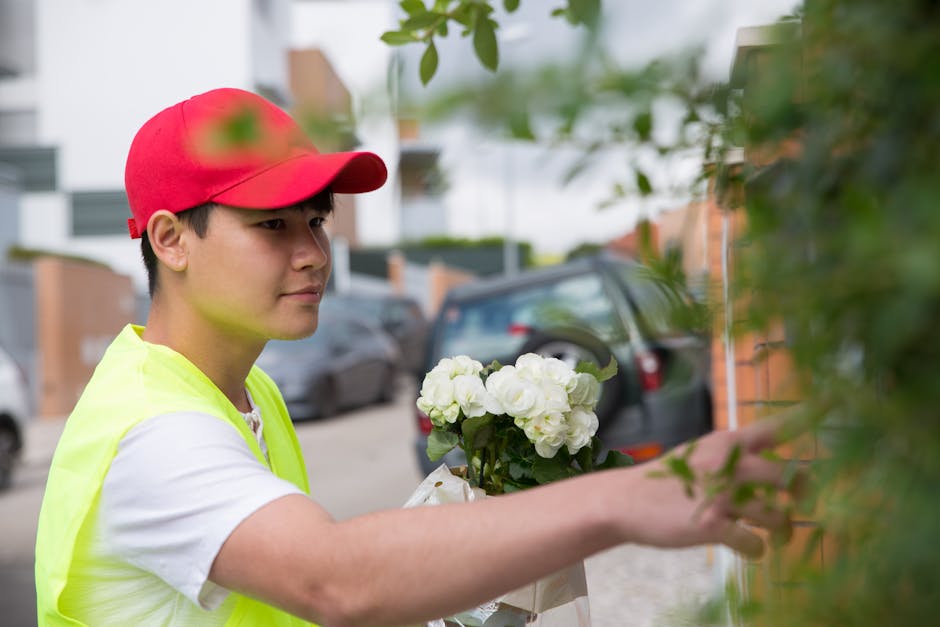 A young male florist wearing a red cap and a fluorescent yellow vest is delivering a fresh floral bouquet consisting of white hydrangeas and other white blooms, wrapped in clear cellophane. He is exte