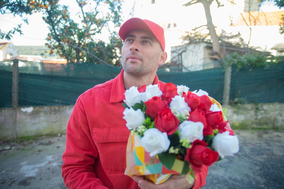 A man dressed in a red uniform and cap is holding a bouquet of fresh flowers, predominantly red and white roses, wrapped in decorative floral paper. He stands outdoors on a paved area with a backgroun