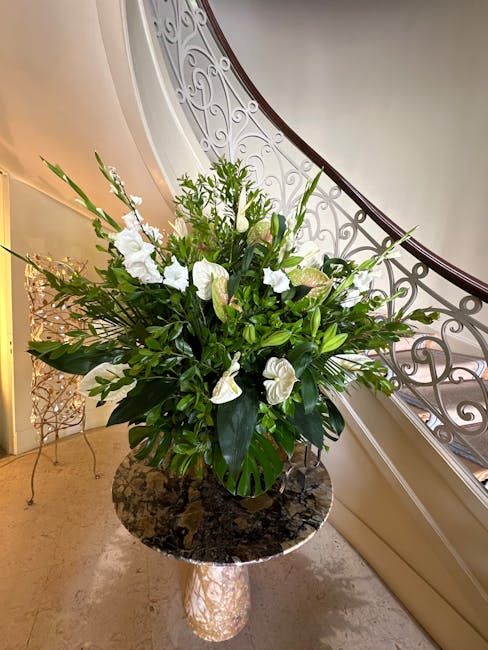A large, elegant floral arrangement featuring white lilies and white anthuriums, accented by lush green foliage, arranged in a black vase. The bouquet is placed on a polished, round marble table with 