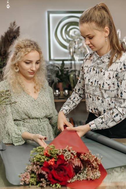 Two women are engaged in a floral arrangement process indoors, with a large bouquet featuring vibrant red roses, pink and green foliage, and small white accent flowers wrapped in red paper in front of