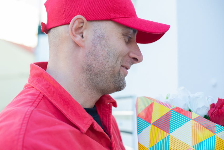 A delivery person wearing a red cap and matching red jacket is holding a floral gift wrapped in colorful, geometric-patterned paper with yellow, red, blue, and green shapes. The flowers inside include