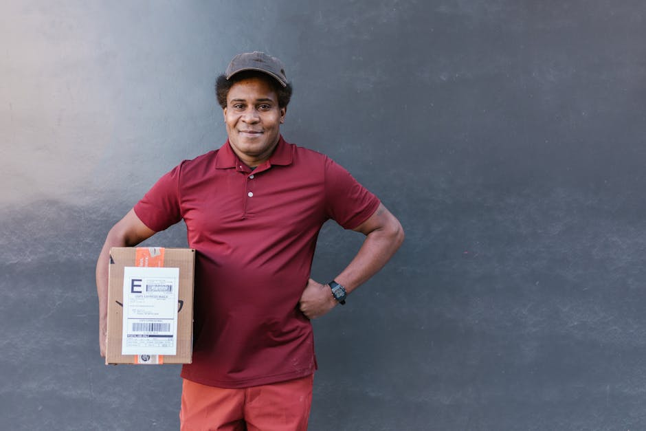 A smiling delivery person with medium skin tone, wearing a maroon polo shirt, matching shorts, and a dark cap, stands against a plain gray wall. He holds a small cardboard parcel with a shipping label