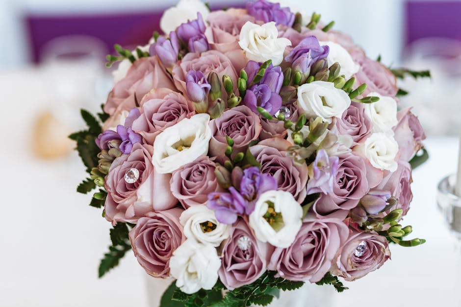 A close-up of a delicate floral bouquet arranged with soft pink roses, white lisianthus, and purple freesias, accented with small green buds and foliage. The flowers are fresh, with slightly ruffled p